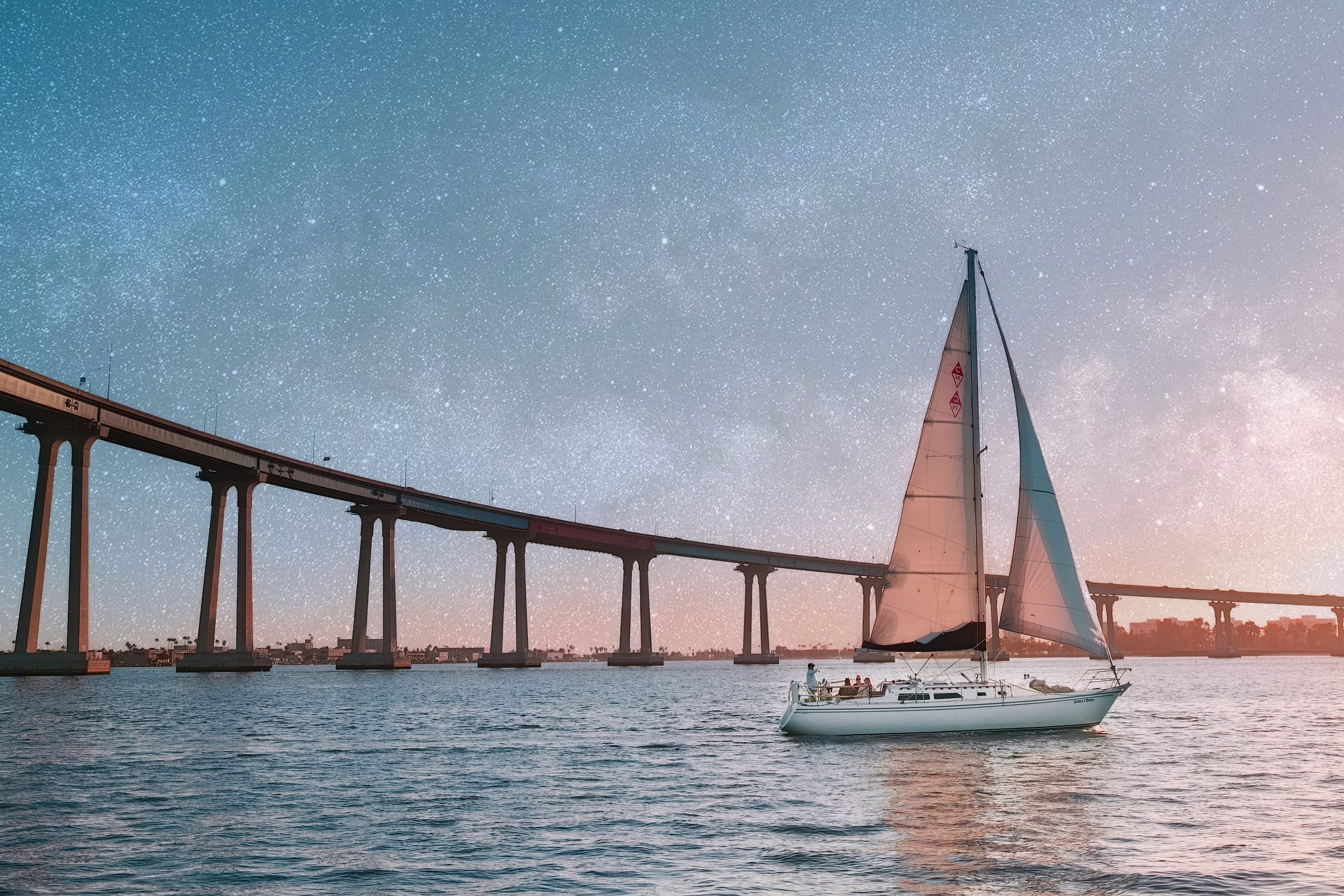 A sailboat passing beneath the Coronado Bridge in San Diego under a dramatic star-filled sky.